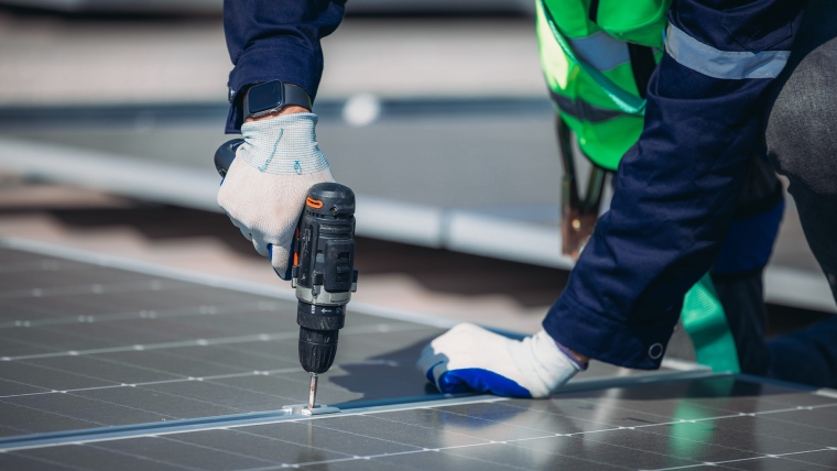 A worker in safety gear is using a drill to secure solar panels on a rooftop under bright sunlight. The setting showcases a modern approach to renewable energy solutions.