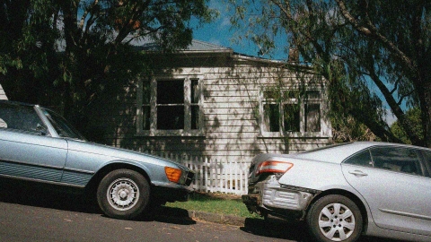 Two cars are parked outside in front of a house in New Zealand. One car is damaged.