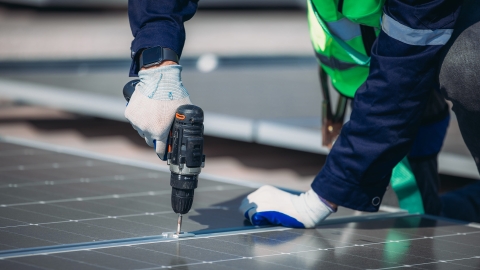 A worker in safety gear is using a drill to secure solar panels on a rooftop under bright sunlight. The setting showcases a modern approach to renewable energy solutions.